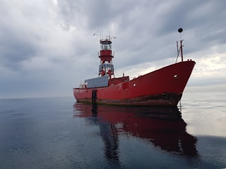 A large red ship with a tower stands motionless on a calm body of water under a cloudy sky. The ship's reflection is visible on the water's surface, and the vessel appears to have solar panels on its side.