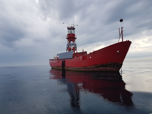 A large red ship with a tower stands motionless on a calm body of water under a cloudy sky. The ship's reflection is visible on the water's surface, and the vessel appears to have solar panels on its side.