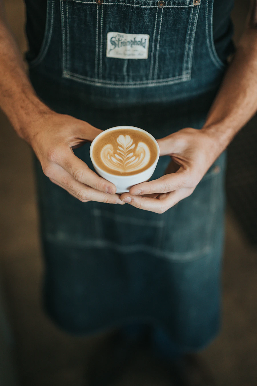A man in a worn denim apron holding a small white cup of coffee in both hands, framed from the chest down, the poured rosetta looking straight up out of the cup.