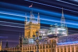 A long exposure shot captures the iconic Palace of Westminster at night, with streaks of blue and white light creating dynamic lines across the sky above the illuminated historic architecture. The Union Jack flag is visible fluttering at the top of the building.
