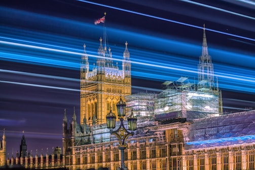 A long exposure shot captures the iconic Palace of Westminster at night, with streaks of blue and white light creating dynamic lines across the sky above the illuminated historic architecture. The Union Jack flag is visible fluttering at the top of the building.