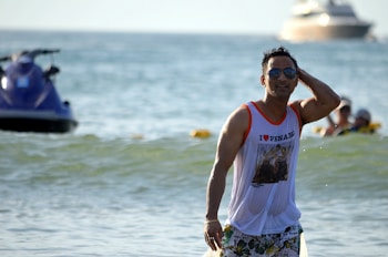 A man wearing sunglasses and a white tank top with 'I Love Penang' printed on it, walks through shallow water at the beach. He appears relaxed and is touching his head with one hand. In the background, there's a large boat and a jet ski in the water.