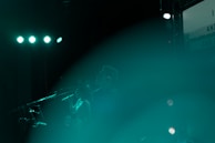 A stage performance featuring silhouetted figures of musicians with microphones, illuminated by teal lights and a hazy atmosphere.