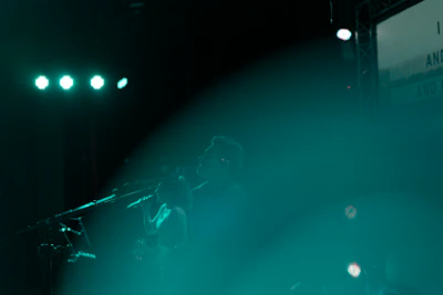 A stage performance featuring silhouetted figures of musicians with microphones, illuminated by teal lights and a hazy atmosphere.