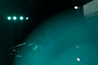 A stage performance featuring silhouetted figures of musicians with microphones, illuminated by teal lights and a hazy atmosphere.