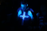 A dimly lit environment with a person wearing a shirt featuring a large cross symbol. The individual is partially illuminated by blue light, creating a moody atmosphere. Other people are faintly visible in the background.