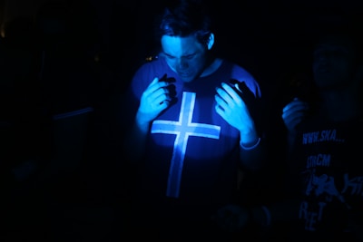 A dimly lit environment with a person wearing a shirt featuring a large cross symbol. The individual is partially illuminated by blue light, creating a moody atmosphere. Other people are faintly visible in the background.