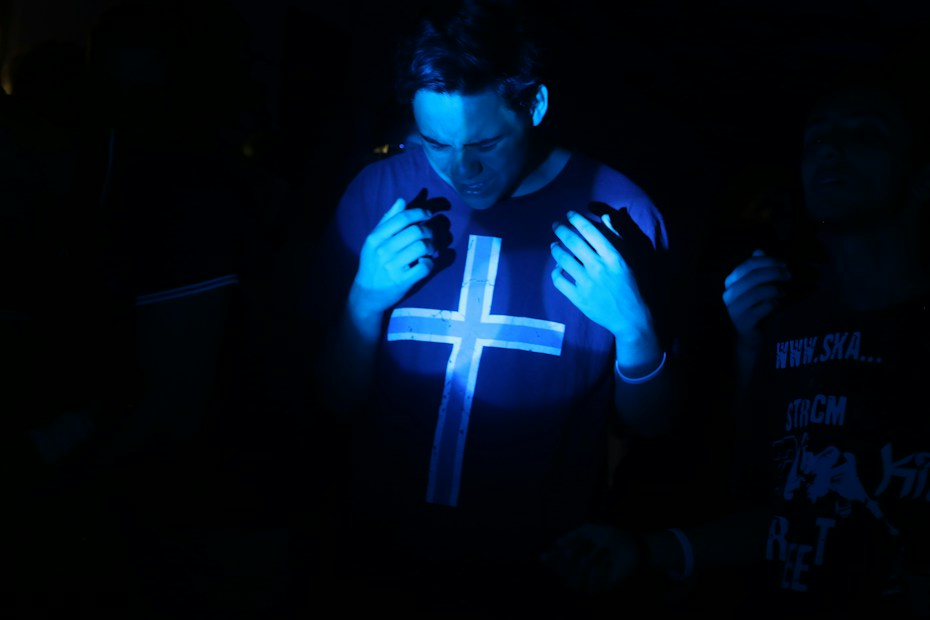 A dimly lit environment with a person wearing a shirt featuring a large cross symbol. The individual is partially illuminated by blue light, creating a moody atmosphere. Other people are faintly visible in the background.