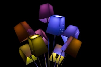 A cluster of colorful, geometric lampshades is arranged closely against a dark background. The lampshades vary in color, with hues of purple, blue, yellow, and orange, creating a vivid contrast with the black backdrop. The light emanating from the lamps is soft, adding an artistic touch to the overall composition.