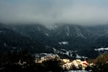 A quaint village nestled at the foot of dark, forested hills, partially covered by snow, with heavy, low-lying clouds above. The village features a small cluster of buildings with red and brown rooftops that contrast with the white snow and dark trees. Warm sunlight illuminates parts of the village, adding a touch of brightness.