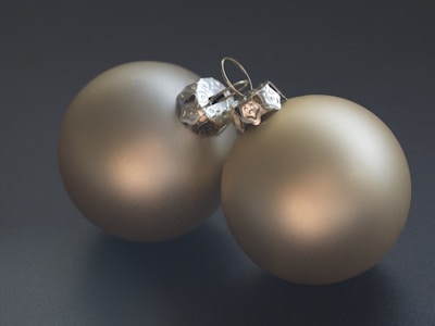Two matte gold Christmas ornaments are lying adjacent on a dark surface. The lighting creates a soft shine on the ornaments, highlighting their smooth, metallic texture. The silver hooks on top have an intricate design.