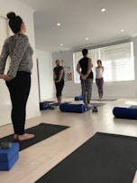 A group of people are standing on yoga mats in a well-lit room with white walls and wooden flooring. They appear to be in a yoga or meditation class. Each person is standing in a relaxed posture with their hands behind their backs. The room has several yoga props including bolsters and blocks. Natural light is streaming in through the large window blinds on one side of the room.
