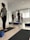 A group of people are standing on yoga mats in a well-lit room with white walls and wooden flooring. They appear to be in a yoga or meditation class. Each person is standing in a relaxed posture with their hands behind their backs. The room has several yoga props including bolsters and blocks. Natural light is streaming in through the large window blinds on one side of the room.