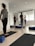 A group of people are standing on yoga mats in a well-lit room with white walls and wooden flooring. They appear to be in a yoga or meditation class. Each person is standing in a relaxed posture with their hands behind their backs. The room has several yoga props including bolsters and blocks. Natural light is streaming in through the large window blinds on one side of the room.