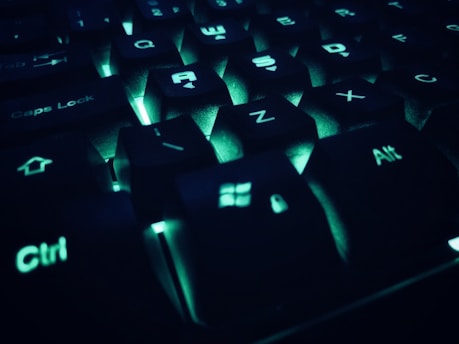 A close-up view of a computer keyboard with keys illuminated by soft, greenish-blue backlighting. The keys are in sharp focus, revealing common function keys like Caps Lock, Ctrl, and Alt, along with letter keys such as A, S, and Z. The lighting creates a futuristic and tech-oriented ambiance.
