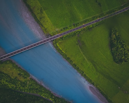 An aerial view of a bridge spanning across a serene blue river, surrounded by lush green fields. The scene captures the intersection of nature and infrastructure, showcasing a harmonious blend of natural beauty and man-made structure.