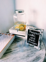 A glass jar with a metal lid sits on a white wooden crate, containing lemon and lime slices submerged in water. Nearby, a framed chalkboard sign reads 'Purified water and fresh oranges.' Several small clear plastic cups are scattered on a round wooden table with a rustic texture.
