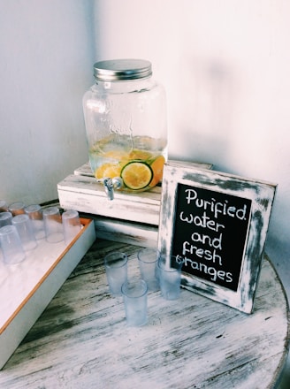 A glass jar with a metal lid sits on a white wooden crate, containing lemon and lime slices submerged in water. Nearby, a framed chalkboard sign reads 'Purified water and fresh oranges.' Several small clear plastic cups are scattered on a round wooden table with a rustic texture.