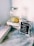 A glass jar with a metal lid sits on a white wooden crate, containing lemon and lime slices submerged in water. Nearby, a framed chalkboard sign reads 'Purified water and fresh oranges.' Several small clear plastic cups are scattered on a round wooden table with a rustic texture.