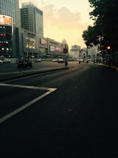 Urban scene with a wide road lined by tall modern buildings and illuminated billboards. Vehicles, including cars and motorcycles, are traveling in both directions. The sky has a soft, yellowish hue, suggesting it is either sunrise or sunset. Trees line one side of the street, adding a touch of greenery.