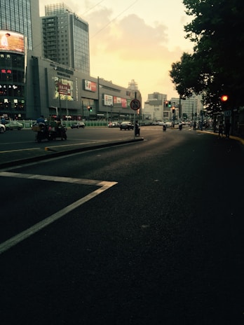 Urban scene with a wide road lined by tall modern buildings and illuminated billboards. Vehicles, including cars and motorcycles, are traveling in both directions. The sky has a soft, yellowish hue, suggesting it is either sunrise or sunset. Trees line one side of the street, adding a touch of greenery.