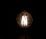 A single illuminated incandescent light bulb against a dark background, with visible glowing filaments inside a round glass bulb.