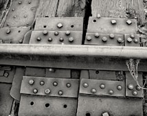 A close-up view of a railway track section showing metal plates, bolts, and wooden railroad ties. The image highlights the textures and details of rusty bolts, weathered wood, and the rough surface of the metal rail. Some weeds and small plants are visible growing around the ties, adding a natural element to the industrial scene.