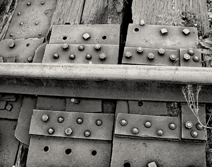 A close-up view of a railway track section showing metal plates, bolts, and wooden railroad ties. The image highlights the textures and details of rusty bolts, weathered wood, and the rough surface of the metal rail. Some weeds and small plants are visible growing around the ties, adding a natural element to the industrial scene.