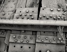 A close-up view of a railway track section showing metal plates, bolts, and wooden railroad ties. The image highlights the textures and details of rusty bolts, weathered wood, and the rough surface of the metal rail. Some weeds and small plants are visible growing around the ties, adding a natural element to the industrial scene.