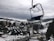 A snowy mountainous landscape with a cluster of buildings, including cabins and larger structures. Pine trees are scattered throughout the area, and a ski lift chair is prominently hanging in the foreground. Overcast skies create a moody atmosphere, and the surrounding hills are partially covered in snow.
