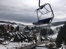 A snowy mountainous landscape with a cluster of buildings, including cabins and larger structures. Pine trees are scattered throughout the area, and a ski lift chair is prominently hanging in the foreground. Overcast skies create a moody atmosphere, and the surrounding hills are partially covered in snow.