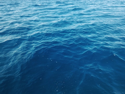 Waves of the ocean extend into the horizon, creating a rhythmic pattern of dark blue shades. The water appears calm with subtle ripples across the surface.