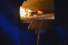 An ancient-looking stone oven with glowing embers and flames inside, casting a warm, golden light. The oven's opening reveals a wooden paddle used for baking, with intricate grain patterns visible on the surface. The surrounding area is dimly lit with a bluish hue, providing a stark contrast to the fiery interior.