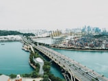 A sprawling urban landscape featuring a large port with numerous shipping containers and cranes. A bridge extends over a body of water, linking green, landscaped areas to the port. Skyscrapers dominate the distant skyline, contrasting with the lush greenery on the left.