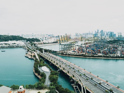 A sprawling urban landscape featuring a large port with numerous shipping containers and cranes. A bridge extends over a body of water, linking green, landscaped areas to the port. Skyscrapers dominate the distant skyline, contrasting with the lush greenery on the left.