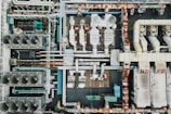 An aerial view of an industrial rooftop filled with various machinery and ductwork. The image features complex networks of pipes, large vents, and possibly HVAC units. The surfaces show signs of rust and wear, indicating age or exposure to the elements.