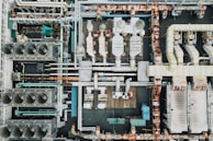 An aerial view of an industrial rooftop filled with various machinery and ductwork. The image features complex networks of pipes, large vents, and possibly HVAC units. The surfaces show signs of rust and wear, indicating age or exposure to the elements.