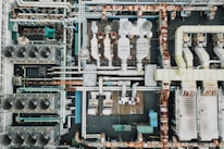 An aerial view of an industrial rooftop filled with various machinery and ductwork. The image features complex networks of pipes, large vents, and possibly HVAC units. The surfaces show signs of rust and wear, indicating age or exposure to the elements.