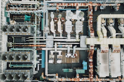 An aerial view of an industrial rooftop filled with various machinery and ductwork. The image features complex networks of pipes, large vents, and possibly HVAC units. The surfaces show signs of rust and wear, indicating age or exposure to the elements.
