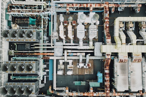 An aerial view of an industrial rooftop filled with various machinery and ductwork. The image features complex networks of pipes, large vents, and possibly HVAC units. The surfaces show signs of rust and wear, indicating age or exposure to the elements.