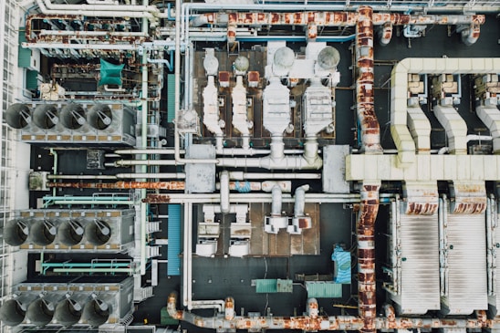 An aerial view of an industrial rooftop filled with various machinery and ductwork. The image features complex networks of pipes, large vents, and possibly HVAC units. The surfaces show signs of rust and wear, indicating age or exposure to the elements.