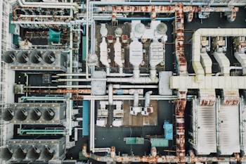 An aerial view of an industrial rooftop filled with various machinery and ductwork. The image features complex networks of pipes, large vents, and possibly HVAC units. The surfaces show signs of rust and wear, indicating age or exposure to the elements.