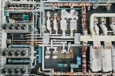 An aerial view of an industrial rooftop filled with various machinery and ductwork. The image features complex networks of pipes, large vents, and possibly HVAC units. The surfaces show signs of rust and wear, indicating age or exposure to the elements.