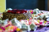 A round chocolate cake topped with a glossy layer of red fruit glaze is surrounded by an assortment of wrapped candies. The cake is decorated with chocolate sprinkles and is placed on a table covered with a light-colored cloth.