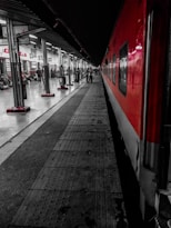 A platform at a railway station with a red and gray train parked alongside. The platform is busy with people walking and sitting. Signs in different languages are visible above various shops and entrances. The lighting is stark, with overhead lights illuminating the space.