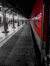 A platform at a railway station with a red and gray train parked alongside. The platform is busy with people walking and sitting. Signs in different languages are visible above various shops and entrances. The lighting is stark, with overhead lights illuminating the space.