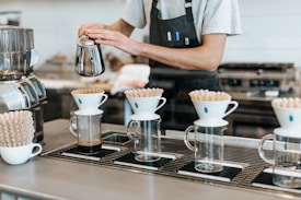 A barista is preparing coffee using a pour-over method. Four glass carafes are lined up with white ceramic drippers on top, each containing a paper filter. Coffee is being poured from a metallic kettle into one of the filters. The barista is wearing a grey shirt with a black apron, and the scene takes place in a clean, modern coffee shop.
