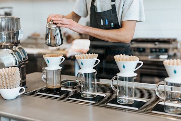 A barista is preparing coffee using a pour-over method. Four glass carafes are lined up with white ceramic drippers on top, each containing a paper filter. Coffee is being poured from a metallic kettle into one of the filters. The barista is wearing a grey shirt with a black apron, and the scene takes place in a clean, modern coffee shop.