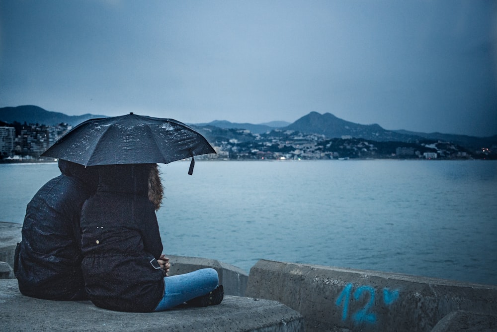 couple holding black umbrella while sitting beside calm water