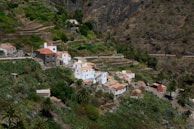 A small, lush rural village nestled within terraced hills and dense vegetation. The village is composed of white and earthy-toned buildings with red-tiled roofs. The landscape surrounding the village is characterized by steep, rocky hillsides and a variety of greenery, including palm trees and other shrubs.